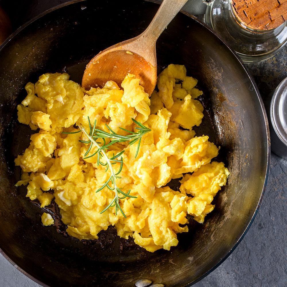 fluffy scrambled eggs in cast iron pan with wooden spatula and rosemary on top